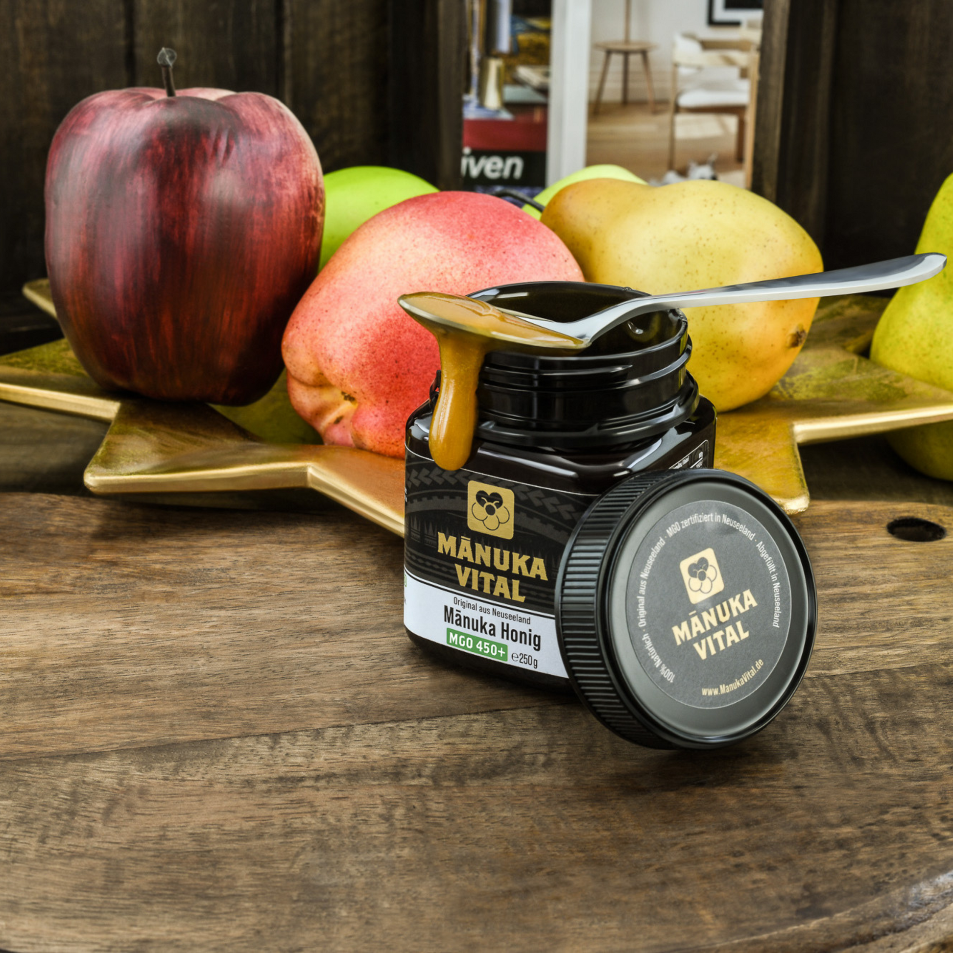 Offenes Glas Manuka Vital Honig mit einem Löffel voll Honig auf dem Glas und Obst im Hintergrund