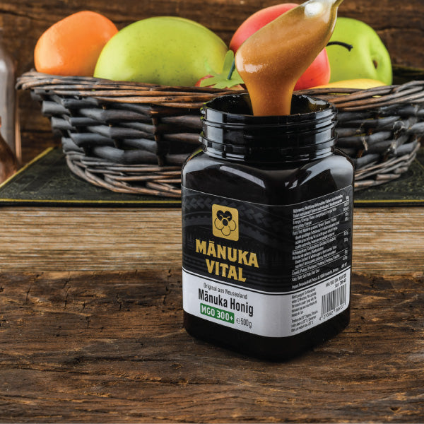 Offenes Glas Manuka Honig MGO 300 – ein Löffel Honig wird entnommen, im Hintergrund frische, gesunde Früchte